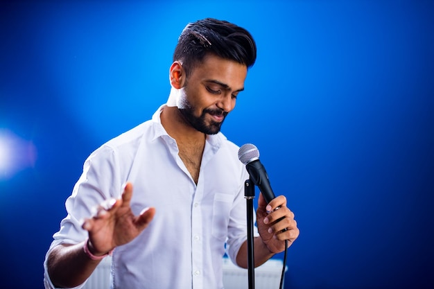 Homme indien en chemise blanche avec barbe chantant en studio d'enregistrement sur fond bleu