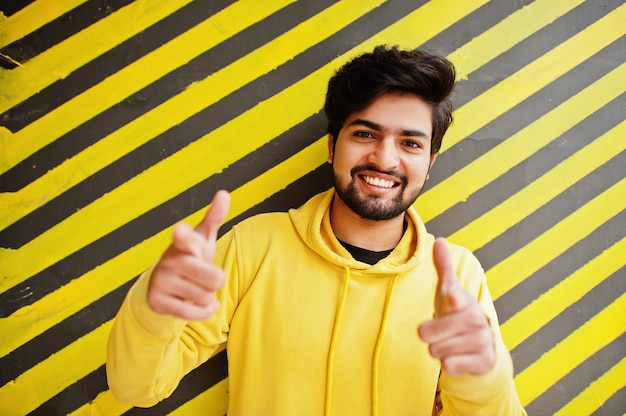 Homme indien jeune hipster urbain dans un sweat-shirt jaune à la mode. Un gars sud-asiatique cool porte un sweat à capuche sur fond rayé, montre les doigts à la caméra.