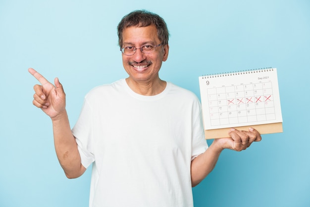 Homme indien senior tenant un calendrier isolé sur fond bleu souriant et pointant de côté, montrant quelque chose dans un espace vide.