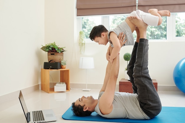 Homme indien souriant allongé sur un tapis de yoga et jouant avec son petit fils qui rit