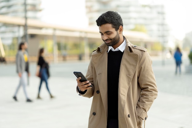 Homme indien souriant tenant un téléphone portable lisant un message texte, communication debout dans une rue urbaine