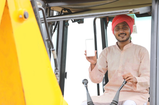 Homme indien travaillant avec des machines de terrassement lourdes et montrant l'écran du smartphone.