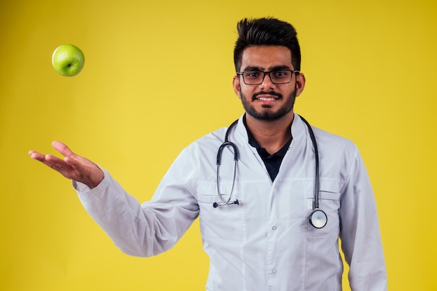 Homme indien en uniforme médical mangeant des pommes sur fond de studio jaune.