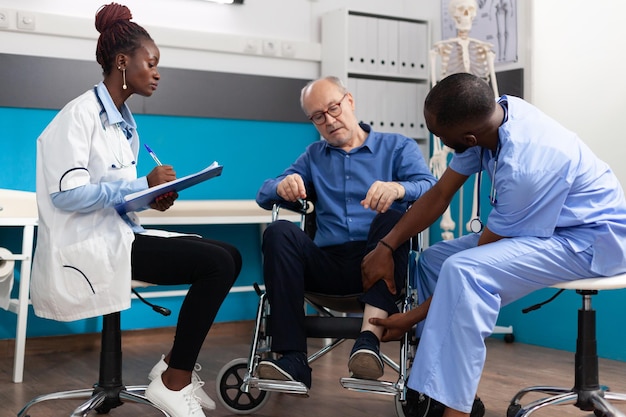 Un homme infirmier vérifie la jambe du genou blessée d'un patient âgé à la retraite pendant que le médecin thérapeute écrit une expertise médicale sur le presse-papiers lors d'un examen clinique au bureau de l'hôpital. Notion de médecine