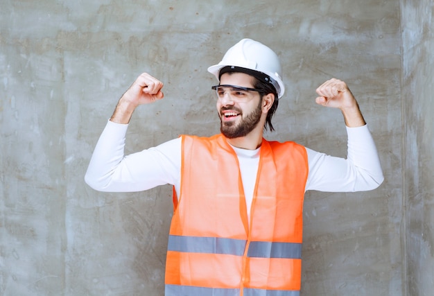 Homme ingénieur en casque blanc et lunettes de protection montrant les muscles de ses bras.