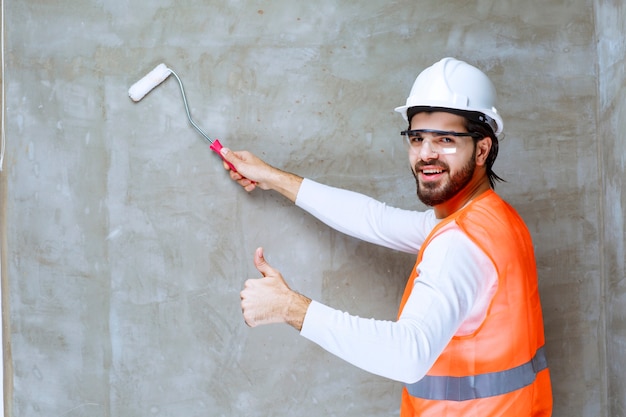 Homme Ingénieur En Casque Blanc Et Lunettes De Protection Peignant Le Mur Avec Un Rouleau De Finition Et Montrant Le Pouce Vers Le Haut.