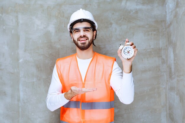 Homme ingénieur en casque blanc et lunettes de protection tenant un réveil.