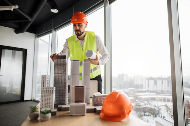 Homme ingénieur en casque et gilet réfléchissant debout près de la table avec la conception de l'architecture de la ville