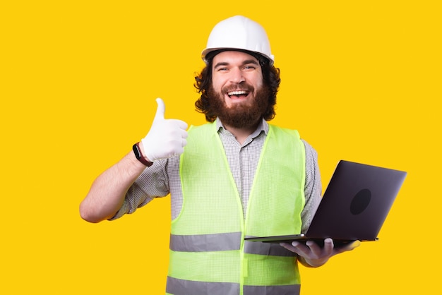 Homme ingénieur gai portant un casque et tenant un ordinateur portable et montrant le pouce vers le haut