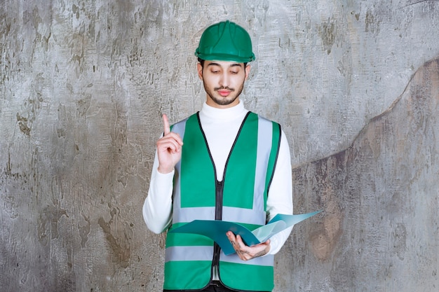 Homme ingénieur en uniforme jaune et casque tenant un dossier bleu et levant la main pour attirer l'attention.