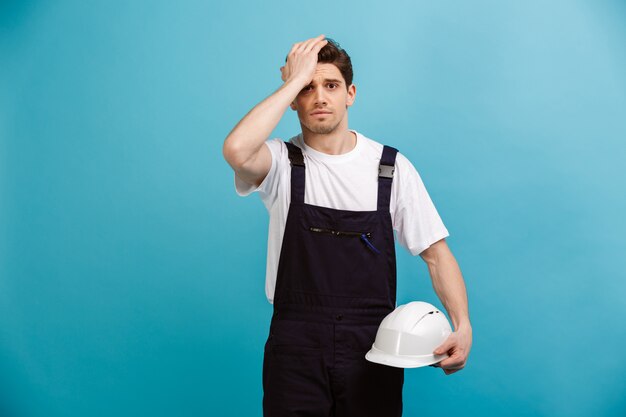 Homme inquiet builder holding casque de protection tout en touchant headf sur mur bleu