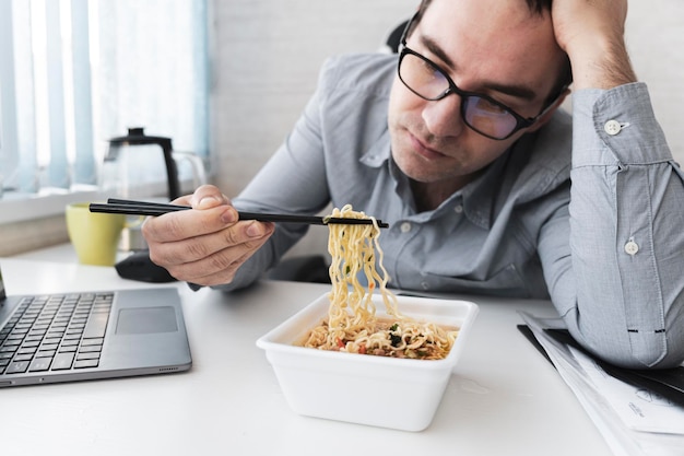 Homme insatisfait mangeant des nouilles instantanées tout en travaillant avec un ordinateur portable au bureau Déjeuner au bureau de la malbouffe insipide