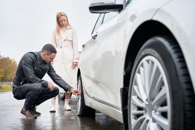 Homme inspectant le pneu endommagé de la voiture blanche de la fille