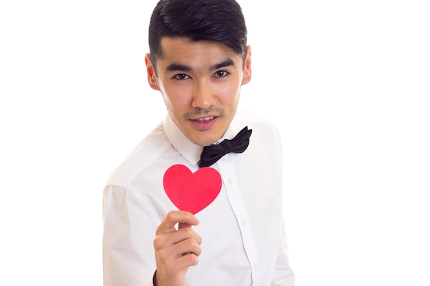 Homme intelligent aux cheveux noirs en T-shirt blanc avec noeud papillon noir tenant un coeur de papier rouge sur fond blanc en studio