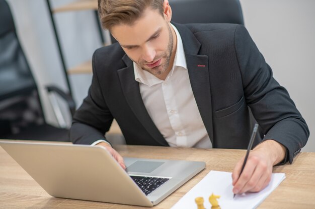 Homme intéressé en costume et chemise blanche assis sur le lieu de travail en face du mouvement d'échecs d'enregistrement d'ordinateur portable