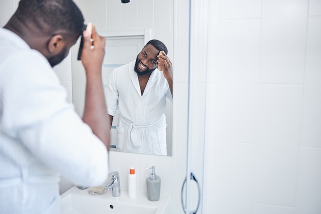 Un homme international ravi et positif se peignant les cheveux tout en regardant son reflet dans le grand miroir