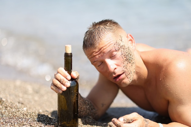 Homme ivre sur la plage avec les yeux fermés tenant une bouteille de vin buvant de l'alcool en chaleur sur la plage