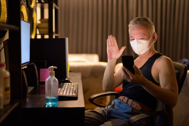 Homme japonais mature avec masque appel vidéo avec téléphone tout en faisant des heures supplémentaires à la maison
