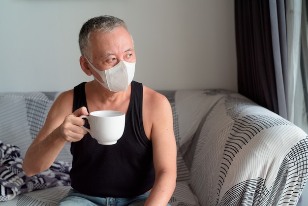 Homme japonais mature avec masque pensant et buvant du café à la maison en quarantaine