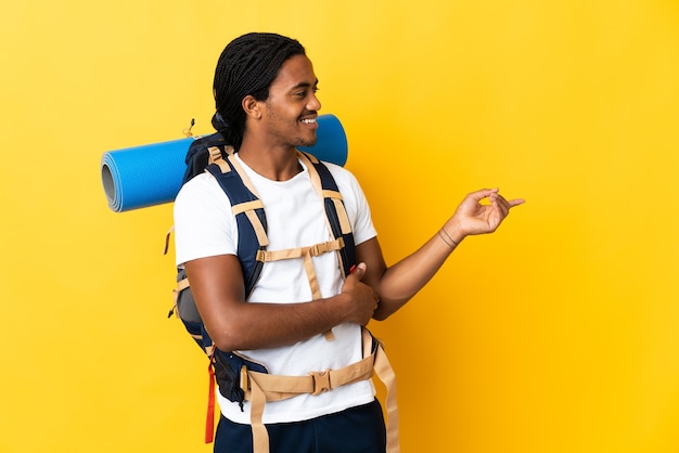 Homme jeune alpiniste avec des tresses avec un gros sac à dos isolé sur fond jaune doigt pointé sur le côté