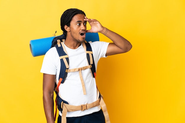 Homme jeune alpiniste avec des tresses avec un gros sac à dos isolé sur fond jaune faisant un geste de surprise tout en regardant sur le côté