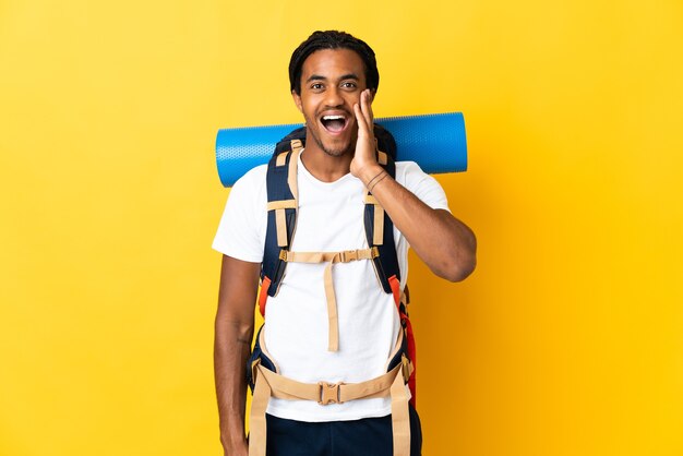 Homme jeune alpiniste avec des tresses avec un gros sac à dos isolé sur un mur jaune avec surprise et expression du visage choqué