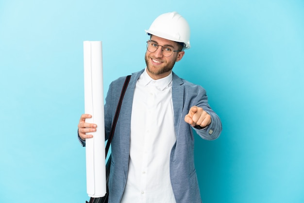 Homme jeune architecte avec casque et tenant des plans sur l'avant pointant isolé avec une expression heureuse