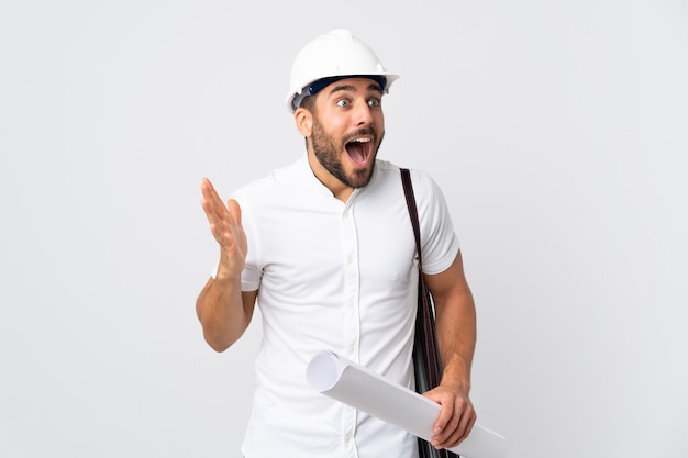 Homme jeune architecte avec casque et tenant des plans isolés sur blanc avec une expression faciale surprise