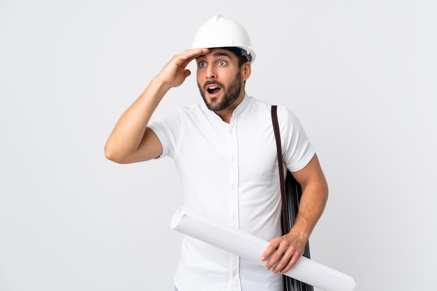 Homme jeune architecte avec casque et tenant des plans isolés sur blanc avec une expression de surprise tout en regardant côté