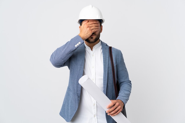 Homme jeune architecte avec casque et tenant des plans isolés sur fond blanc couvrant les yeux par les mains. Je ne veux pas voir quelque chose