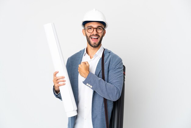 Homme jeune architecte avec casque et tenant des plans isolés sur un mur blanc célébrant une victoire