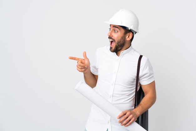 Homme jeune architecte avec casque et tenant des plans isolés sur un mur blanc pointant le doigt sur le côté et présentant un produit