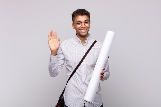 Homme jeune architecte souriant joyeusement et gaiement, en agitant la main, en vous accueillant et en vous saluant, ou en vous disant au revoir