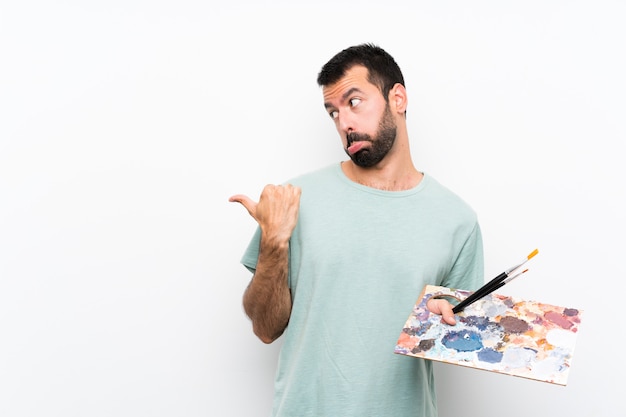 Homme jeune artiste tenant une palette sur fond isolé malheureux et pointant vers le côté