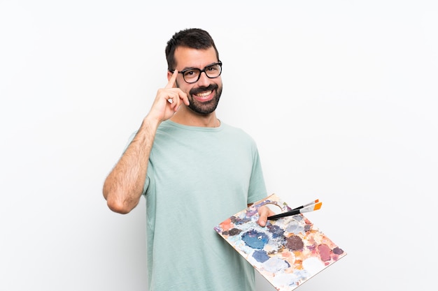 Homme jeune artiste tenant une palette sur un mur isolé avec des lunettes et heureux