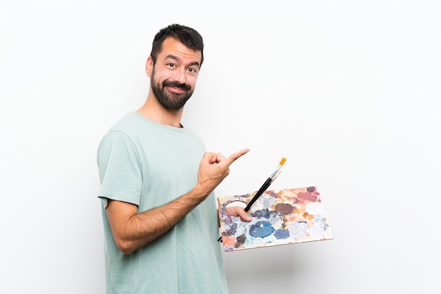 Homme jeune artiste tenant une palette sur un mur isolé pointant sur le côté pour présenter un produit