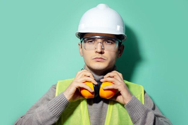 homme jeune constructeur, ingénieur, touchant son casque de construction sur le cou, portant un casque de sécurité et des lunettes sur le mur de couleur aqua menthe