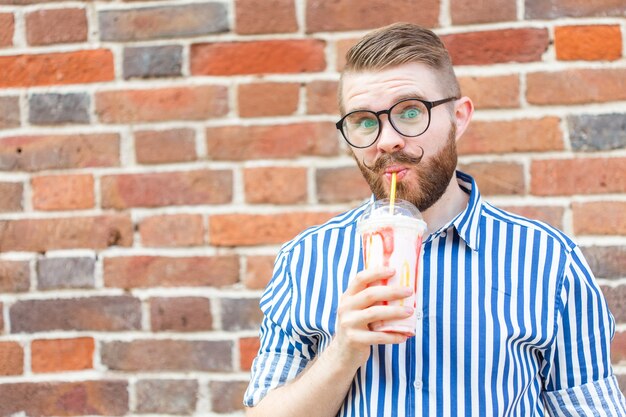 Homme jeune hipster positif flou avec barbe et moustache dans des verres tenant un milkshake aux fruits dans ses mains posant dans la rue près d'un mur de briques avec espace de copie. Concept de promenades d'été.