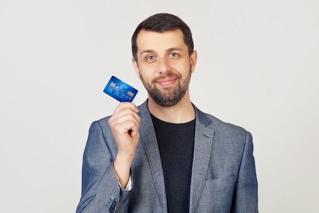 Homme jeune homme d'affaires avec une barbe dans une veste tenant une carte de crédit avec une expression confiante sur un visage intelligent et en pensant sérieusement.