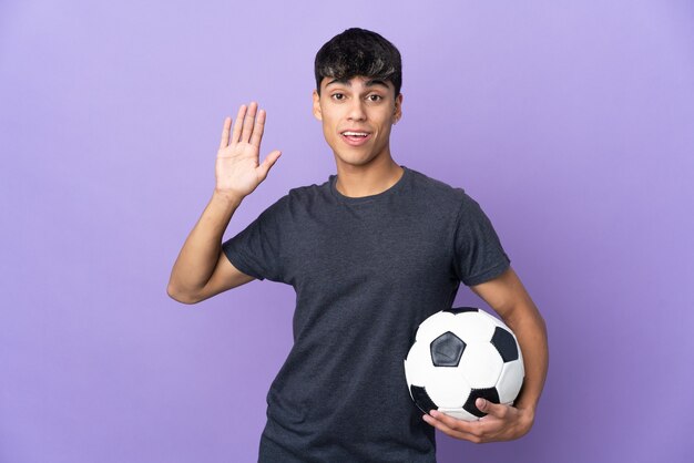 Homme jeune joueur de football sur mur violet isolé saluant avec la main avec une expression heureuse