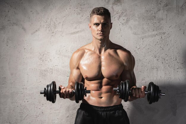 Homme jeune et musclé bodybuilder travaillant avec des haltères contre le mur de béton