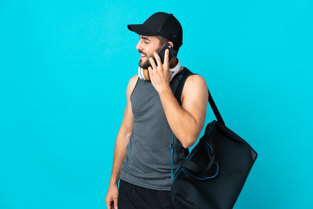Homme jeune sport avec sac de sport isolé sur bleu en gardant une conversation avec le téléphone mobile avec quelqu'un