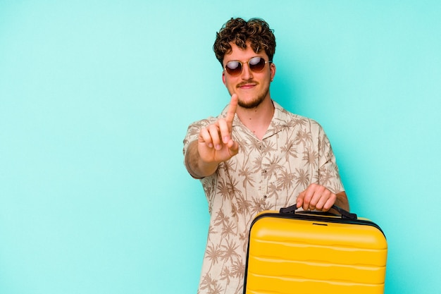 Homme jeune voyageur tenant une valise jaune sur un mur bleu montrant le numéro un avec le doigt.