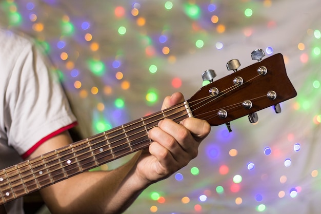 Homme jouant à la guitare acoustique. Gros plan des doigts sur le manche de la guitare contre les lumières de bokeh floues de Noël sur l'arrière-plan.