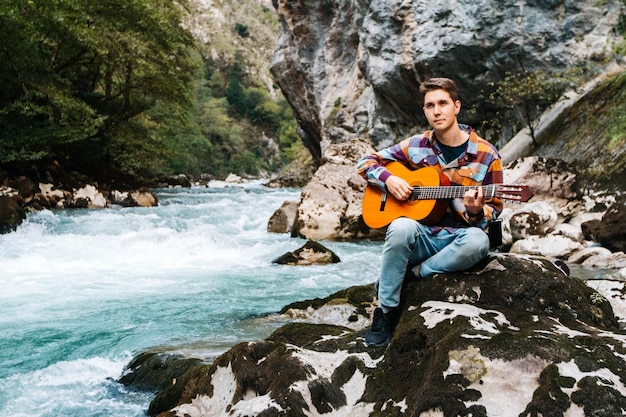 Homme Jouant De La Guitare Assis Sur La Rive D'une Rivière De Montagne Sur Fond De Rochers Et De Forêt