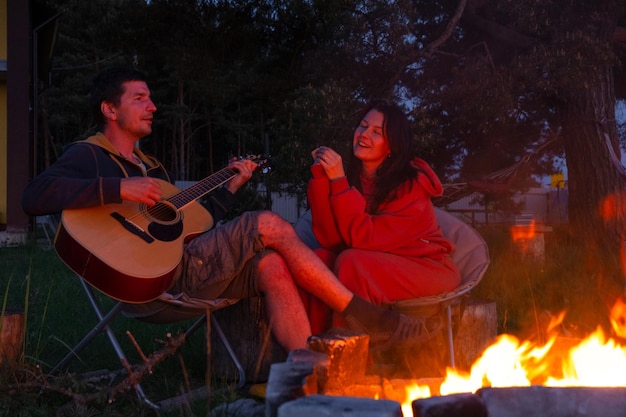 Un homme joue de la guitare une femme écoute et chante un couple amoureux est assis près du feu de camp en plein air dans la cour de la maison sur des chaises de camping une soirée romantique