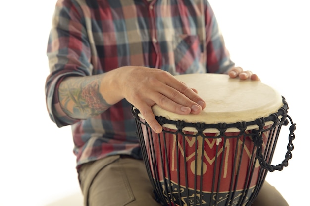 L'homme joue des percussions de darbouka à tambour ethnique, gros plan musicien isolé sur fond de studio blanc. Mains masculines tapant djembé, bongo en rythme. Instruments de musique faits à la main, son de la culture mondiale.