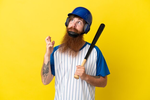 Homme de joueur de baseball rousse avec casque et batte isolé sur fond jaune avec les doigts croisés et souhaitant le meilleur