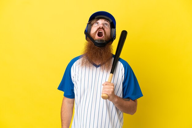 Homme de joueur de baseball rousse avec casque et batte isolé sur fond jaune en levant et avec une expression surprise