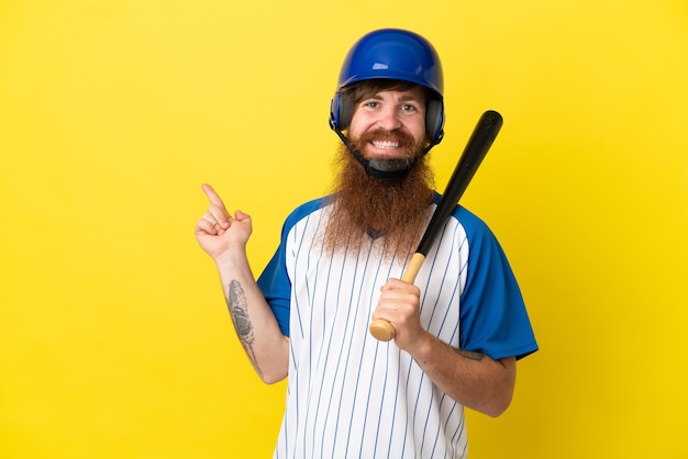 Homme de joueur de baseball rousse avec casque et batte isolé sur fond jaune, pointant le doigt sur le côté